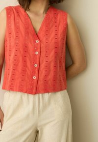 Blouse sans manches corail avec un col en V, présentant des découpes circulaires, cinq boutons sur le devant, et une coupe ample, associée à un pantalon léger en lin.