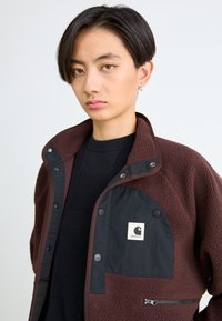 Persona joven con pelo corto y oscuro que lleva una chaqueta de forro polar marrón con panel negro y el logo de Carhartt sobre un suéter negro, fondo neutro.
