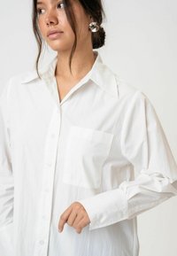 Fehér, hosszú ujjú, gombos ing hegyes gallérral, egyetlen mellzsebbel és sima textúrával. Az ujjak enyhén redőzöttek.