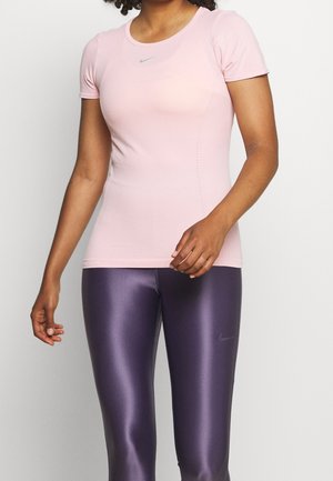 Femme portant un haut moulant rose clair à manches courtes et des leggings violets brillants, debout devant un fond clair uni.