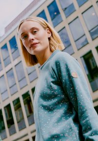 Felpa verde menta con sottili pattern circolari rosa, scollatura a costine e dettagli del logo color tan sulla manica. Texture liscia, vestibilità casual.