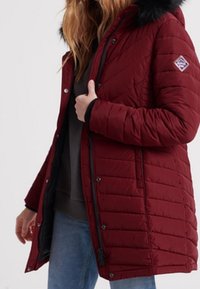 Quiltet bordeaux parka-jakke med en faux fur hætte, der har trykknapper, lynlås og kontrasterende sort foer.