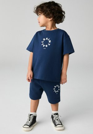 Set di t-shirt e pantaloni corti in cotone blu navy con dettaglio del logo circolare bianco. Abbinato a sneaker nere con lacci bianchi e suole testurizzate.