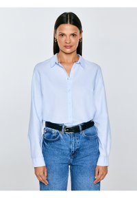 Sieviete ar taisniem tumšiem matiem valkā gaiši zilu krekliņu ar pogām, ieliktu augsta vidukļa zilās džinsās ar melnu jostu, stāvot pret balto fonu.