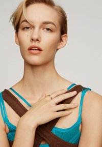 El modelo lleva una prenda de imitación cuero marrón con un diseño acolchado sobre una camiseta sin mangas turquesa. Un anillo dorado en forma de zigzag adorna su mano.