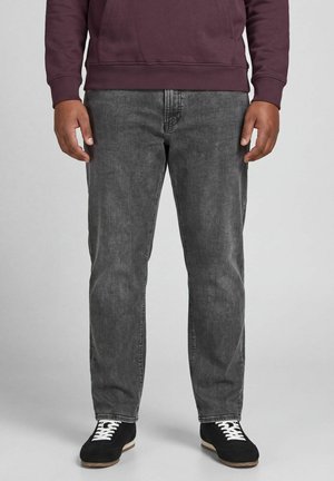 Homme portant un jean en denim gris, des baskets noires avec des lacets blancs et un sweat-shirt violet foncé, debout sur un fond gris.
