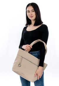 Otse tumedate juustega naine, kes kannab musta üllatust ja siniseid teksapükse, hoiab käes beežikat nahast käekotti, millel on ees tõmblukk ja lai rihm.