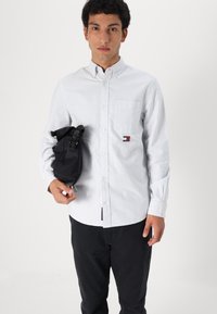 Button-up shirt in white with light blue stripes, long sleeves, pocket on left side, worn with black bag in hand, paired with black pants.