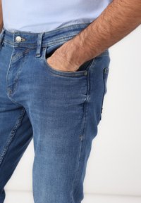Jeans en denim bleu clair avec un design classique à cinq poches. Présentent des coutures contrastées et une fermeture à bouton en métal. Texture douce.