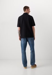 Sort polo skjorte med korte ærmer, parret med blå jeans. Synlig bagstikning og let tekstur på skjorten. Brune sko fuldender outfittet.