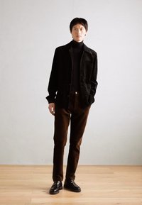 Sort fløjljakke, tætsiddende turtleneck, brune fløjlbukser og sorte loafers; modellen står på trægulv foran en ensfarvet væg.