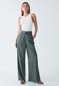 Pantalon large vert en tissu lisse, avec taille haute, passants pour ceinture et poches latérales, associé à un débardeur blanc ajusté.