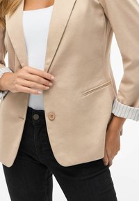 Beige blazer met een enkele knoop, minimalistisch ontwerp en geplooide textuur. De mouwen hebben een wit en grijs gestreept patroon. Draagt over zwarte jeans.