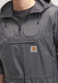 Czarna i szara lekka kurtka z kapturem, z przednią kieszenią na zamek, ściągaczem u dołu oraz wyraźnym pomarańczowym logo Carhartt.