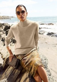 Frau in beigem Rollkragenpullover, gemustertem Rock, großen Sonnenbrille und mit einer geflochtenen Clutch, sitzt auf Steinen am Ozeanrand.