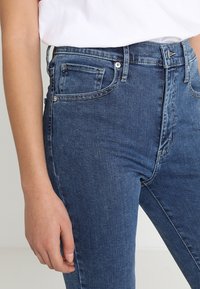 Jeans de denim azul de cintura alta con un diseño clásico de cinco bolsillos, que presentan un cierre delantero con botón y un ligero desgaste en la tela.