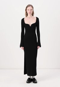 Abito lungo a maniche nere in maglia con scollatura a barca, spacco laterale e un dettaglio decorativo sul petto. Tessuto morbido e design aderente.