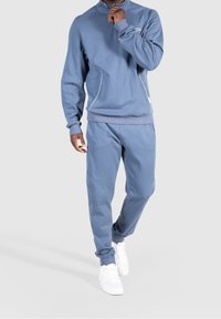Modrý polyesterový teplákový komplet s vysokým límcem, kapsou na zip a bílým prýmkem. Doplněný bílými teniskami pro dokonalý vzhled.