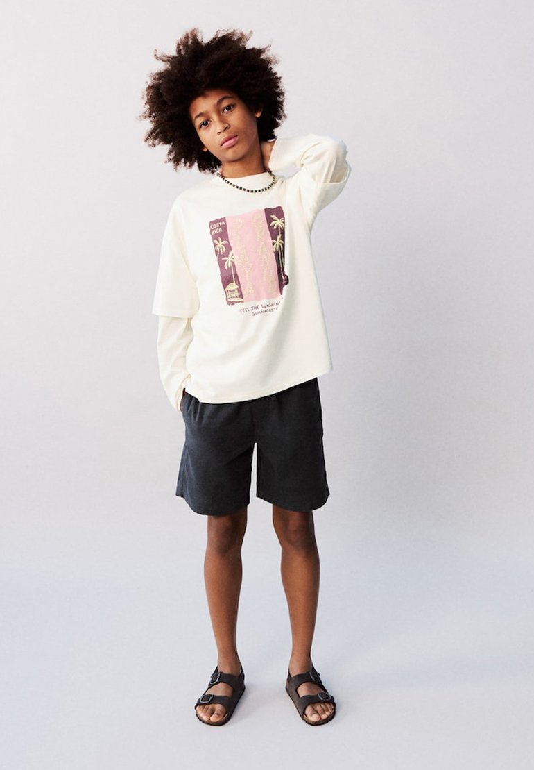 Enfant aux cheveux bouclés portant un t-shirt blanc à manches longues avec un motif graphique, un short noir et des sandales noires, debout avec une main derrière la nuque.