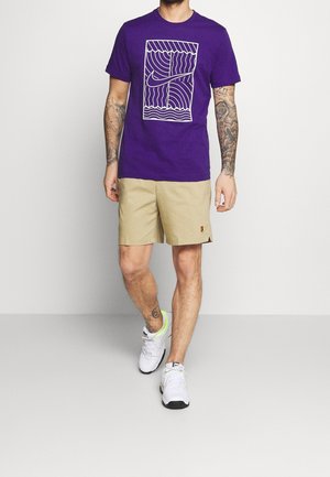 Muž v fialovém tričku s grafikou, béžových šortkách a bílých teniskách, stojící před jednoduchým světlým pozadím, s viditelnými tetováními na rukou a nohách.