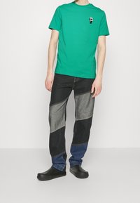 Grøn kortærmet t-shirt med et lille grafisk design på brystet, parret med farveblokerede denimbukser i sort, grå og blå, samt sorte slip-on sko.