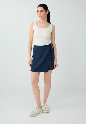 Frau mit dunklen Haaren in einem weißen ärmellosen Top, einem marineblauen Wickelrock und weißen Sneakers steht vor einem schlichten, hellen Hintergrund.