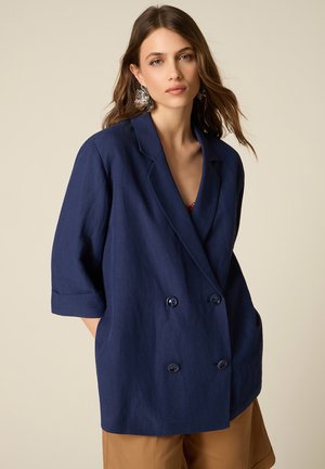 Donna con lunghi capelli castani che indossa grandi orecchini d'argento, blazer doppiopetto blu navy e pantaloni beige su sfondo beige.