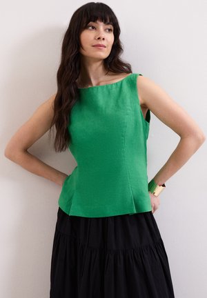 Mujer con cabello largo y oscuro, que lleva una blusa verde sin mangas y una falda negra, de pie con las manos en las caderas frente a una pared lisa.