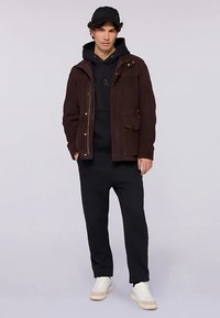 Brązowa kurtka materiałowa z zatrzaskami, kapturem i bocznymi kieszeniami, noszona na czarnym kapturze i czarnych spodniach dresowych, zestawiona z białymi sneakersami.