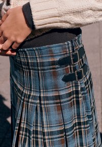 Spódnica w kratę w kolorze niebieskim, zielonym i brązowym z plisami; posiada czarne skórzane klamry w pasie; miękka w dotyku i o konstrukcyjnym kroju.