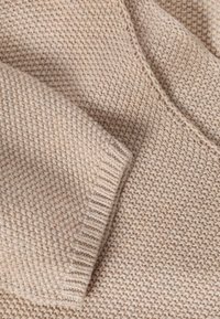 Gros plan d'un tissu tricoté beige montrant un motif de point texturé et le détail du poignet d'une manche longue.