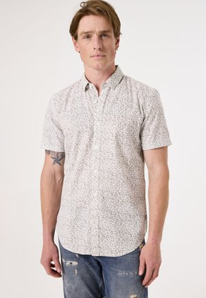 Chemise à manches courtes, à boutons, fabriquée en tissu léger, présentant une base blanche avec un délicat motif de feuilles noires. Portée avec un jean bleu déchiré.