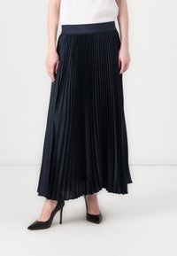 Marineblå plisseret nederdel med en glat tekstur, bred elastisk talje, gulvlangt design, parret med sorte spidse hæle.