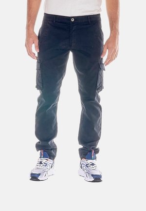 Mann trägt dunkle Cargohosen und weiße blaue Sneakers, steht mit entspannten Händen an den Seiten vor einem weißen Hintergrund.