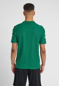 Grøn polo shirt med klassisk krave, korte ærmer og hvide chevron mønstre på skuldrene. Lavet af tekstureret stof.