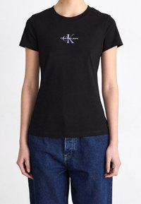 Person, der ein schwarzes Calvin Klein Jeans T-Shirt und blaue Jeans trägt, steht mit entspanntem Arm entlang der Seiten.
