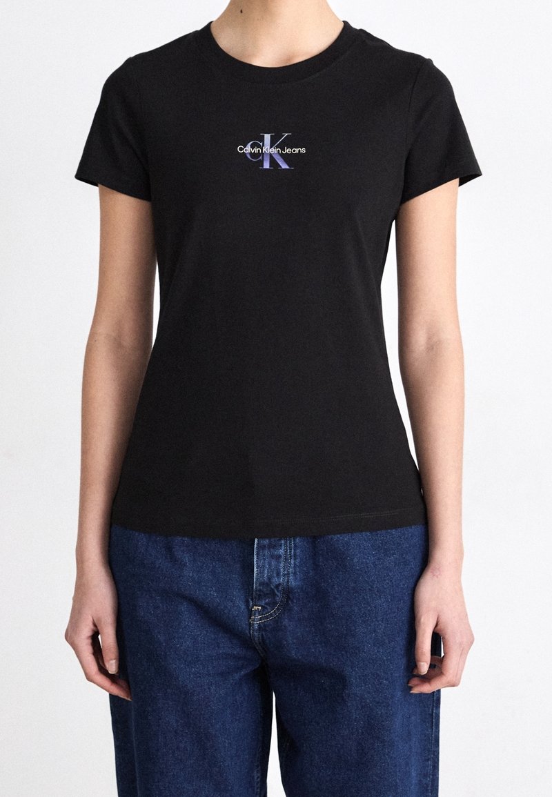 Person, der ein schwarzes Calvin Klein Jeans T-Shirt und blaue Jeans trägt, steht mit entspanntem Arm entlang der Seiten.