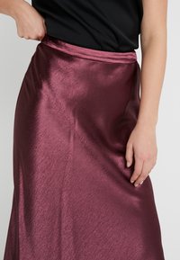 Bordowa satynowa spódnica o gładkiej, błyszczącej fakturze, z dopasowaną talią i płynną sylwetką. Ręka spoczywa na spódnicy.
