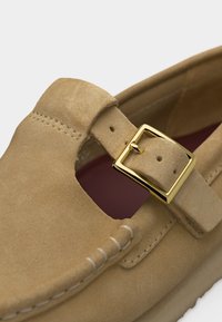 Hellbrauner Wildlederschuh mit goldener Schnallriemen und sichtbaren Nähten entlang vorne und seitlich, zeigt einen Teil der Innensohle.