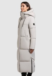 Long puffer coat in light gray, featuring a hood, zippered side pockets, and stitched detailing for insulation. Smooth texture.