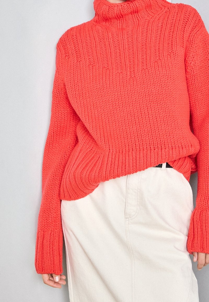 Leuchtender korallenfarbener Rollkragenpullover aus grobem Strickstoff mit gerippter Textur, kombiniert mit einem hellbeigen Rock.