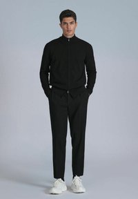 Veste zippée noire et pantalon tailleur en tissu texturé, associés à des baskets blanches. Le design est minimaliste avec une coupe moderne.