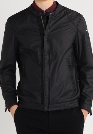Mann trägt eine schwarze gesteppte Jacke mit Reißverschluss und Druckknöpfen, darunter ein burgunderfarbenes Hemd, die Hände in den Taschen, gezeigt vom Hals bis zur Hüfte.