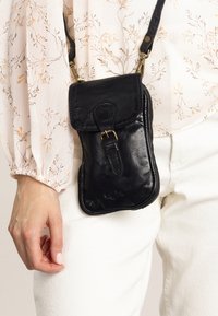 Sac à bandoulière en cuir noir avec un rabat, ferrures en laiton et une poche zippée. Texture lisse et forme rectangulaire.