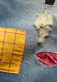 Denim z frędzlem, z dziurą, żółtą łatą w czerwony wzór kratki oraz czerwoną łatą z białymi motywami.