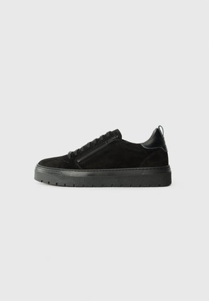Black suede sneaker with a textured leather accent, rounded shape, lace-up closure, and side zipper detail. Thick rubber sole with grooves.