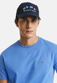 Cappello della marina con bandiere ricamate e la scritta "GANT USA"; indossato con una maglietta blu a collo tondo con un piccolo logo. Tessuto liscio, design informale.