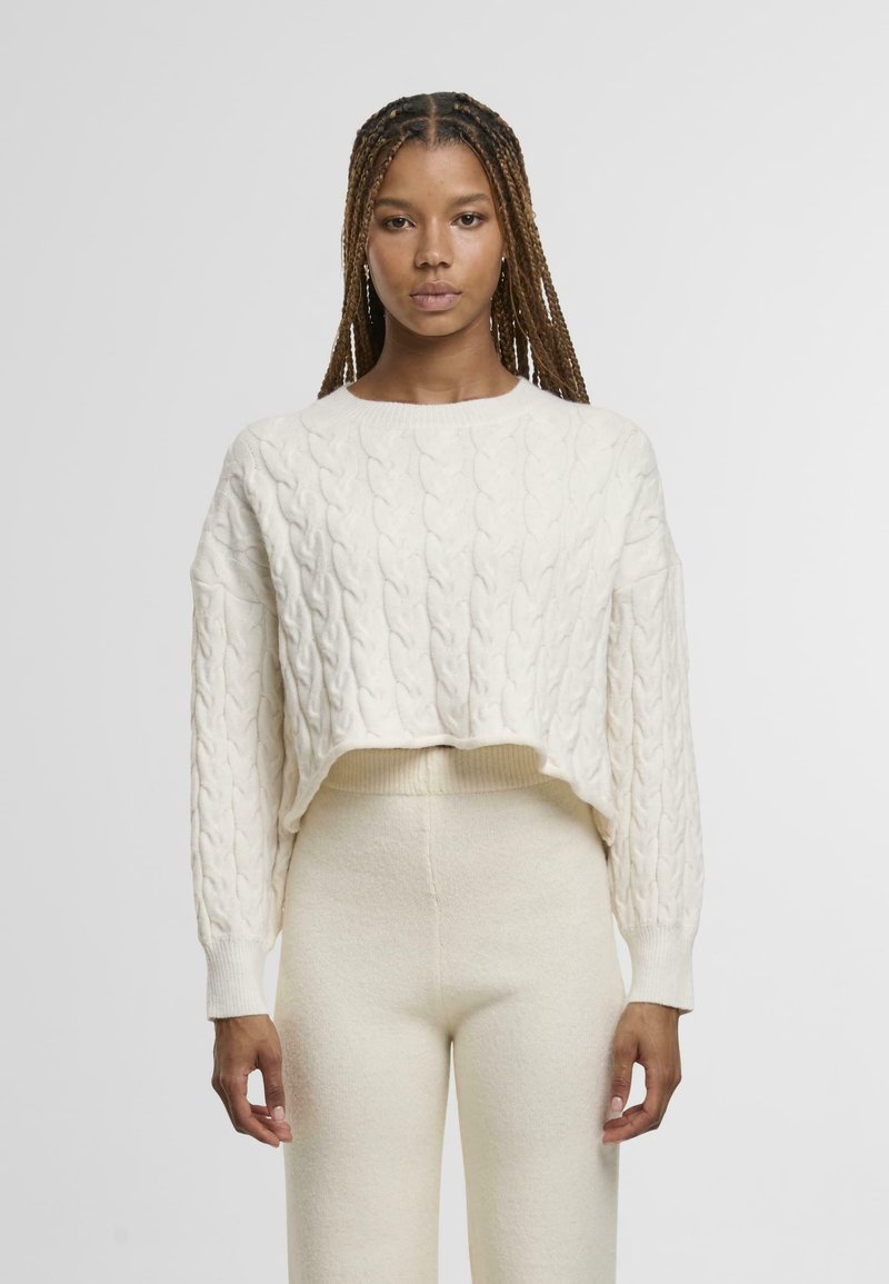 Cream-colored cropped cable-knit sweater with long sleeves and ribbed hem, paired with matching high-waisted cream trousers.