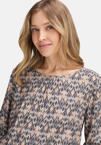 Blusa stampata in grigio, beige e arancione con motivo geometrico. Tessuto leggero con scollatura a barchetta e dettagli plissettati sulle spalle.