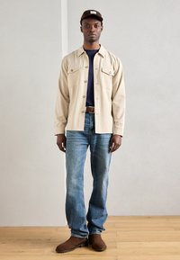 Μπέιζ πουκάμισο με κουμπιά και τσέπες στο στήθος, μπλε μπλούζα από κάτω, μπλε τζιν και καφέ δερμάτινα μποτάκια, ολοκληρωμένο με ένα καφέ καπέλο.